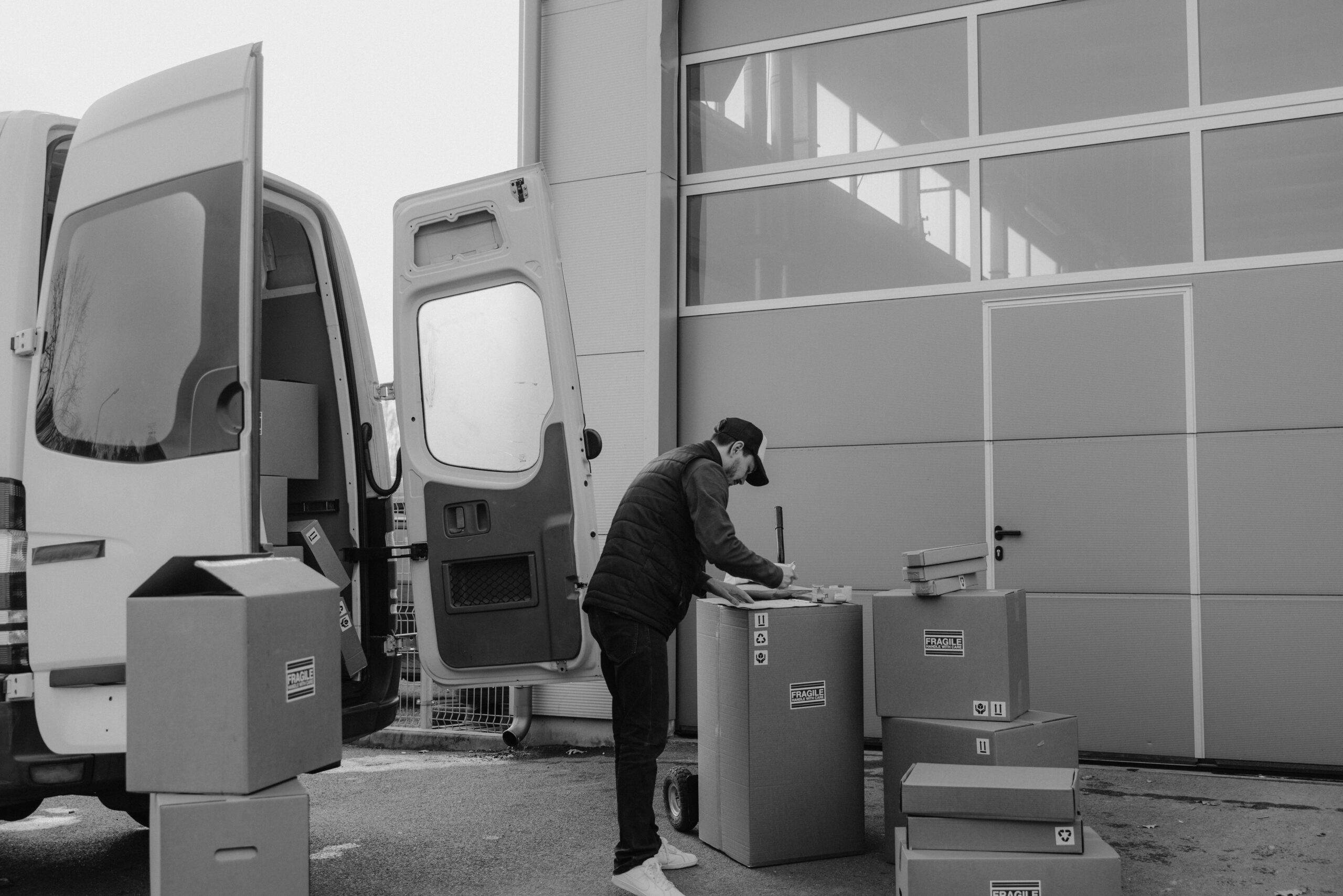 Monochrome image of a courier managing packages outside a warehouse, illustrating logistics workflow.
